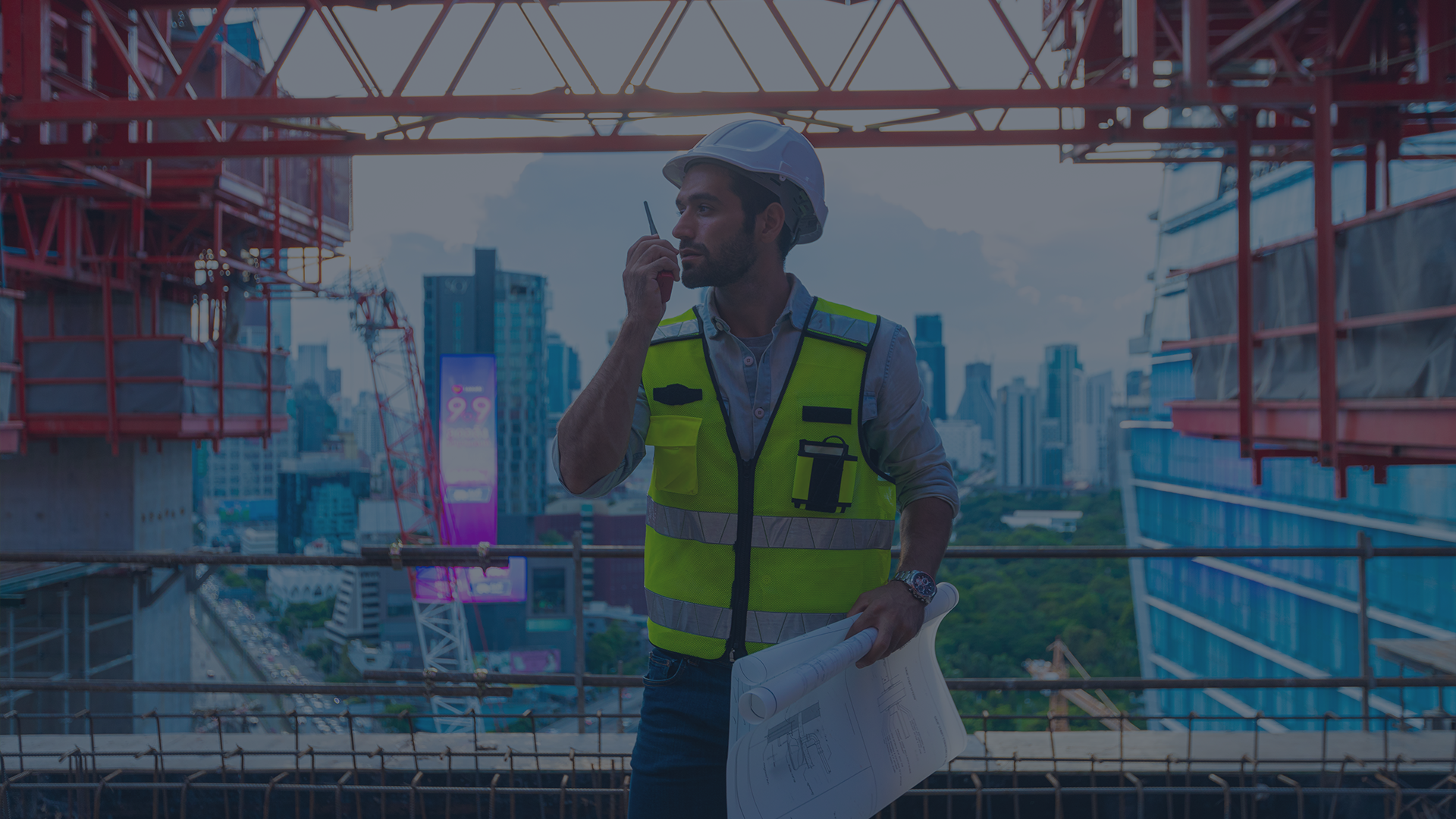 A site supervisor in a hard hat and hi-vis vest coordinates by two-way radio on a New York high-rise under construction, blueprints in hand and a tower crane and city skyline behind him, on a DOB-regulated enterprise jobsite.