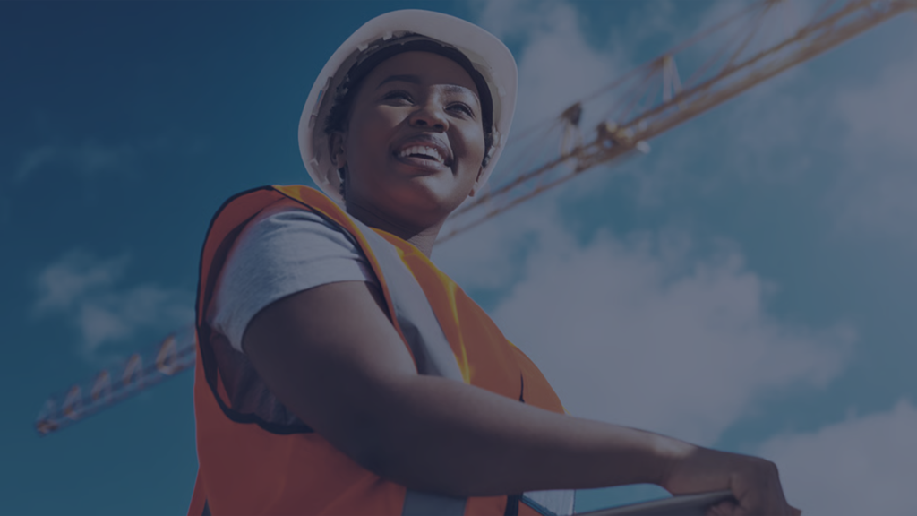 Smiling construction worker in hard hat and hi-vis vest on a jobsite with tower cranes against a blue sky, representing predictive safety management